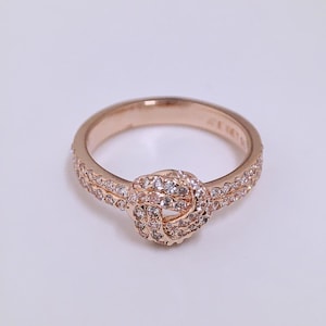 May include: A rose gold ring featuring a central knot design encrusted with small clear stones. The band is split into two parallel rows, each set with a continuous line of small clear stones that extend from the knot to the back of the ring.