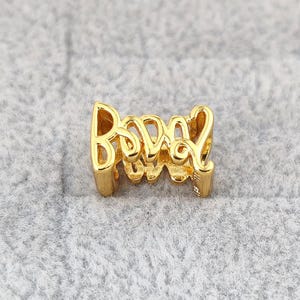 May include: A gold-toned metal charm featuring the word "LOVE" in a stylized, cursive script font. The letters are interconnected, forming a continuous, open-work design with a polished, reflective surface finish that catches the light.