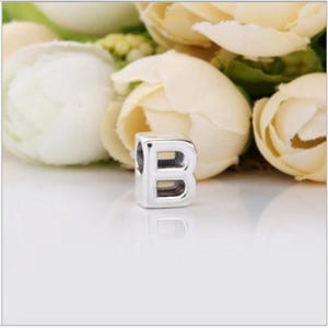 May include: A silver-toned metal bead shaped like the capital letter B. The bead has a smooth, polished finish with a hollow center, designed to be threaded onto a chain or cord. The letter B is clearly embossed on the front surface.