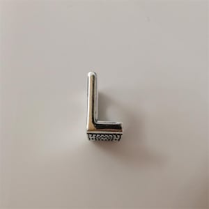 Puede incluir: Un charm de metal plateado con la letra "L". La letra tiene una base texturizada y un acabado pulido. El charm está diseñado para la fabricación de joyas u otros proyectos de artesanía.