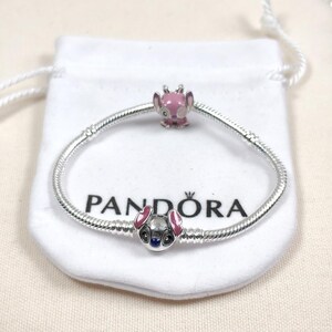 May include: A silver snake chain bracelet featuring two charms. One charm is a pink enamel character head, and the other is a metallic character head with pink ears and dark eyes. The bracelet is displayed on a white fabric pouch with the Pandora logo.