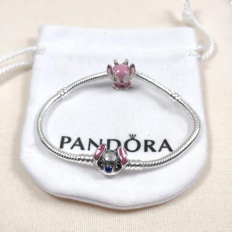 May include: A silver snake chain bracelet featuring two charms. One charm is a pink enamel character head, and the other is a metallic character head with pink ears and dark eyes. The bracelet is displayed on a white fabric pouch with the Pandora logo.