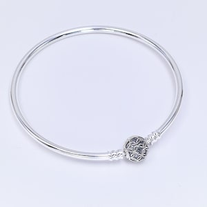 May include: A polished silver bangle bracelet featuring a smooth, circular band and a single spherical charm. The charm displays an intricate, openwork lattice pattern with dark recessed detailing, positioned between two small, ridged silver stoppers.