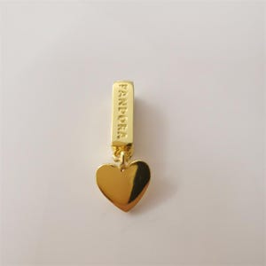 Könnte beinhalten: Ein goldfarbener Charm mit herzförmigem Anhänger. Der Charm hat eine rechteckige Oberseite mit dem eingravierten Wort "PANDORA". Der Herzanhänger ist glatt und reflektierend.