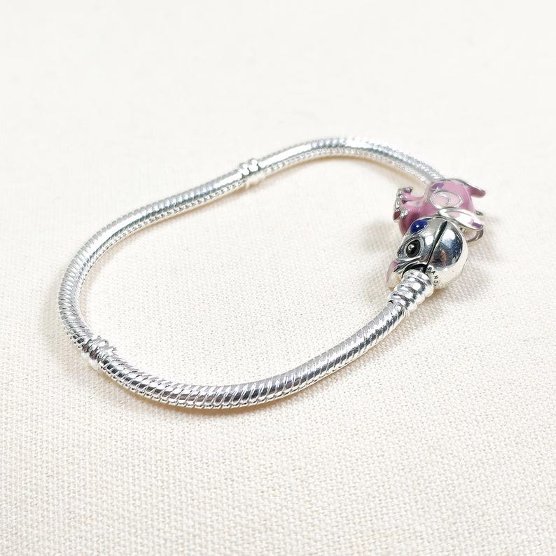 May include: A silver snake chain bracelet featuring a charm shaped like a character with large black eyes, a blue nose, and pink ears. The charm has a metallic finish and is attached to the flexible, textured silver chain with a secure clasp.
