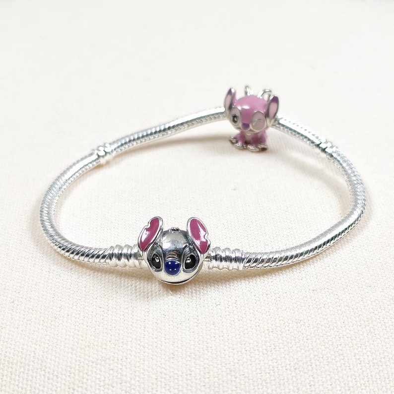 May include: A silver snake chain bracelet featuring two character charms. One charm is a silver-toned Stitch head with pink inner ears and a dark blue nose. The second charm is a pink enamel figure of Angel with large eyes and ears.