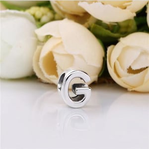 May include: A polished silver-toned metal charm bead shaped like the uppercase letter G. The bead features a smooth, reflective surface with a central opening designed for threading onto a bracelet or necklace chain, showcasing a clean design.