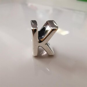 May include: A silver-coloured charm in the shape of the letter "K". The charm has a polished finish and a small hole at the top for stringing. The letter is in a serif font.