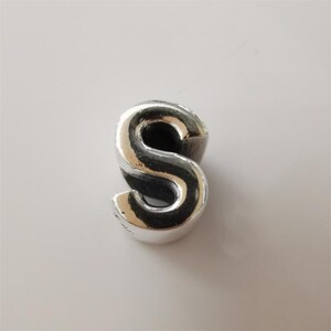 May include: A silver-coloured charm in the shape of the letter "S". The charm has a rounded, three-dimensional design with a smooth, reflective surface. The letter is a capital "S".
