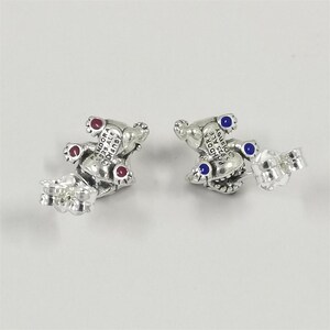 May include: A pair of sterling silver stud earrings shaped like small figures, each featuring the engraved text PANDORA S925 ALE DISNEY. One earring has three red circular accents, while the other earring has three blue circular accents on the feet.