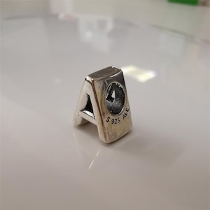 Puede incluir: Un charm de plata con forma de la letra "A". El charm tiene un pequeño diseño circular con el número 4 en su interior. El charm está marcado con "S925 ALE".