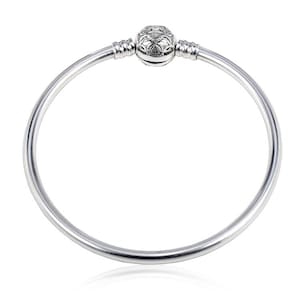May include: A silver bangle bracelet featuring a smooth, circular band and a central spherical clasp. The clasp is decorated with an intricate, raised floral-inspired pattern and is flanked by two ribbed silver segments on the band.