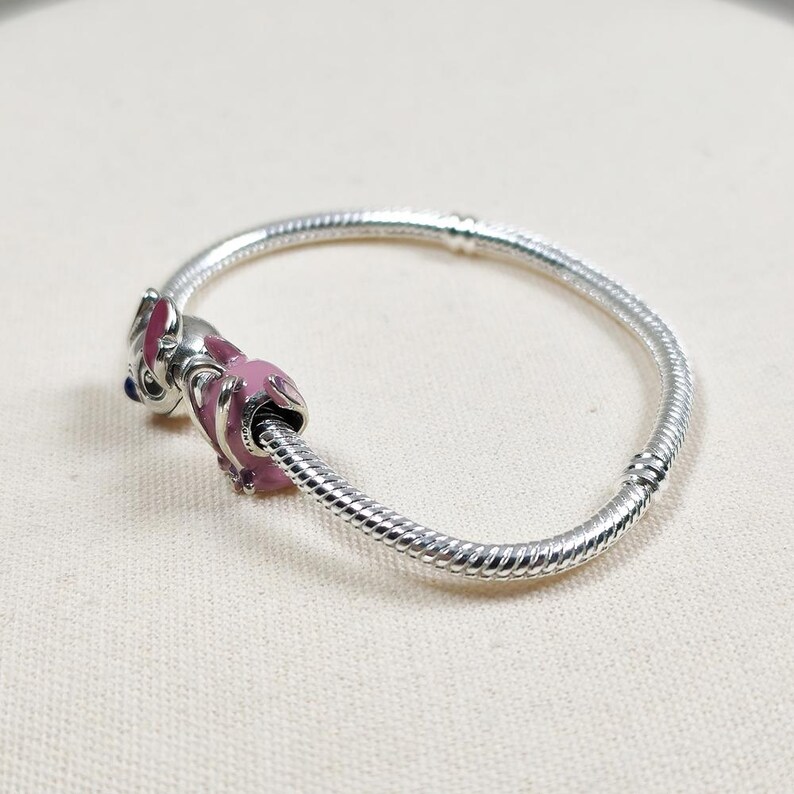 May include: A silver snake chain bracelet featuring a charm shaped like a character with pink enamel detailing and a small blue accent. The bracelet has a smooth, flexible metallic texture and a secure clasp mechanism visible on the side.