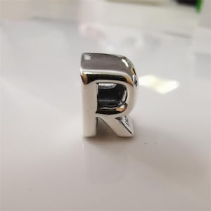 Może przedstawiać: Błyszczący srebrny charms w kształcie litery "R". Charms ma wypolerowaną, odblaskową powierzchnię i wydrążony środek. Litera jest w kroju szeryfowym i stoi prosto.