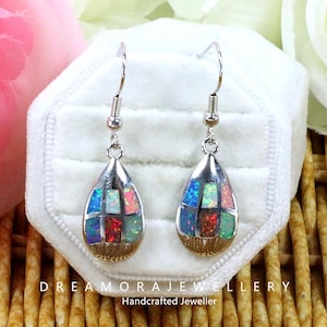 Può includere: Orecchini a goccia in argento con un mosaico di pietre di opale colorate. Gli orecchini hanno una base color oro e sono esposti in un portagioie bianco. Il testo "DREAMORAJEWELLERY Handcrafted Jeweller" è visibile.