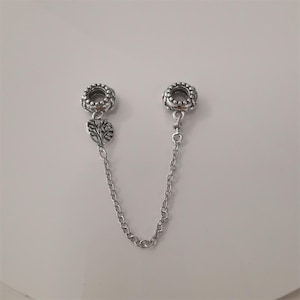 Può includere: Catena di sicurezza per bracciale con ciondoli in argento. Due estremità rotonde e decorate con perline sono collegate da una catena sottile. Un'estremità presenta un ciondolo con un motivo ad albero.