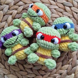 May include: Four handmade crochet turtle toys in green, yellow, and brown. Each turtle has a different coloured mask: orange, blue, red, and purple. The toys are arranged on a woven, textured surface.