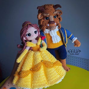 Puede incluir: Muñecos de ganchillo de Bella y la Bestia. Bella lleva un vestido amarillo y sostiene una rosa roja. La Bestia es marrón con una chaqueta azul y amarilla. Los muñecos están hechos a mano.