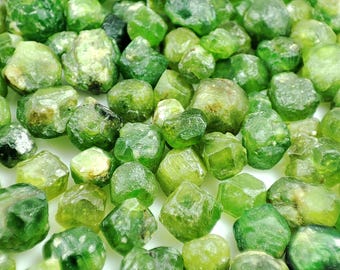 May include: A collection of raw, uncut peridot gemstones. The stones vary in shades of green, from light lime to deep olive, with a natural, unpolished appearance. The gemstones are scattered, showcasing their unique shapes and textures.