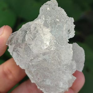 Puede incluir: Un espécimen mineral transparente y translúcido con una estructura cristalina dentada. La superficie presenta numerosas facetas afiladas e irregulares que reflejan la luz, creando una apariencia brillante y gélida con una textura rugosa.