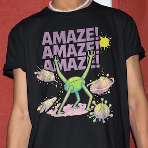 Könnte beinhalten: Schwarzes T-Shirt mit dem Wort "AMAZE!" dreimal in Lila wiederholt, über einer grünen, außerirdisch aussehenden Figur mit erhobenen Armen. Das Design umfasst eine gelbe Sonne und Weltraum-Illustrationen.