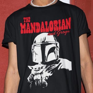 Op de afbeelding: Zwart T-shirt met een witte afbeelding van The Mandalorian en Grogu. De tekst "THE MANDALORIAN and Grogu" is in het rood boven de afbeelding gedrukt. Het ontwerp bevat de helm van de Mandalorian en Grogu.