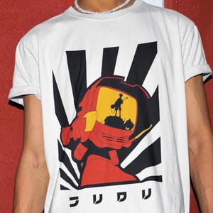 Puede incluir: Camiseta blanca con un diseño gráfico de una cabeza de robot roja, una pantalla amarilla, rayos en blanco y negro y caracteres japoneses. El diseño está centrado en la parte delantera de la camiseta.