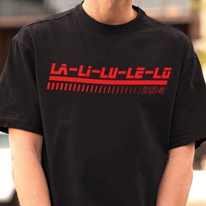 Peut inclure: T-shirt noir avec le texte rouge "LA-LI-LU-LE-LO" et des caractères japonais. Le t-shirt à manches courtes est porté par une personne portant des lunettes de soleil et une casquette noire.