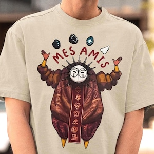 Puede incluir: Camiseta beige de manga corta con un gráfico de una figura enmascarada con una túnica estampada que hace malabarismos con tres objetos. El texto "MES AMIS" está impreso encima de la figura. La persona de la imagen lleva gafas de sol y una gorra negra.