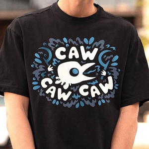 Può includere: T-shirt nero con un design grafico bianco e blu che dice "CAW CAW". Il design presenta un uccello stilizzato con il testo in un carattere giocoso. La maglietta è girocollo e a maniche corte.