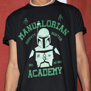 Peut inclure: T-shirt noir avec un motif vert repr&eacute;sentant un casque Mandalorien et les mots "Mandalorian Hunter Academy". Le motif comprend le texte "Bounty" et "Est. 32 BBY."