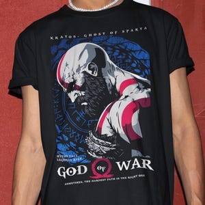 Può includere: T-shirt nero con grafica di Kratos di God of War. Il design include il testo "KRATOS - GHOST OF SPARTA" e "GOD OF WAR". L'immagine ha uno schema di colori blu e rosso, con testo aggiuntivo in basso.