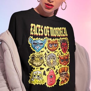 Może przedstawiać: Czarna koszulka z kolorową grafiką przedstawiającą dziewięć postaci z kreskówek i żółtym napisem "FACES OF MORDECAI". Twarze obejmują niebieskiego potwora, niedźwiedzia, diabła, żabę, sowę, demona, kleksa, cyklopa i mężczyznę w okularach.