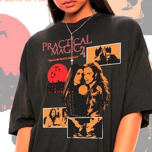 Puede incluir: Camiseta negra con la inscripción "PRACTICAL MAGIC 2" y imágenes de la película. El diseño incluye una luna roja e ilustraciones. La camiseta tiene un aspecto vintage y un corte holgado. El texto "There's No Devil In The Draft" también es visible.