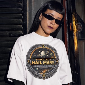 Puede incluir: Camiseta blanca con un gráfico circular que dice "PROJECT HAIL MARY SCIENCE EXCHANGE PROGRAM". El diseño incorpora planetas, estrellas y una combinación de colores dorados y negros. La camiseta la lleva una persona con gafas de sol negras y mangas negras.