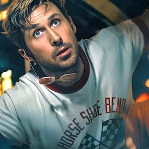 以下が含まれることがあります： 赤い襟と袖が付いた、明るい灰色のTシャツ。Tシャツには「HORSE SHOE BEND」の文字と2つのチェッカーフラッグが描かれています。画像内の人物はメガネをかけています。