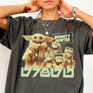 K&ouml;nnte beinhalten: Dunkelgraues Comfort Colors T-Shirt mit einer Grafik von Grogu und anderen Charakteren aus The Mandalorian. Das Design enth&auml;lt den Text "BOUNTY HUNTER" und "GROGU" in gr&uuml;ner Schrift. Das Shirt hat eine lockere Passform.