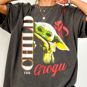 May include: Dark gray Comfort Colors tee featuring a graphic of Grogu, also known as The Child, from the Star Wars series. The design includes the word "CHILD" vertically and "Grogu" in red. The shirt has short sleeves.