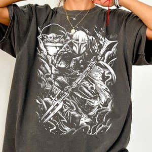Puede incluir: Camiseta gris oscuro Comfort Colors con un gr&aacute;fico blanco de un guerrero mandaloriano y Grogu. El dise&ntilde;o incluye el texto "Comfort Colors Tee (PEPPER)". La camiseta tiene mangas cortas y un corte relajado.