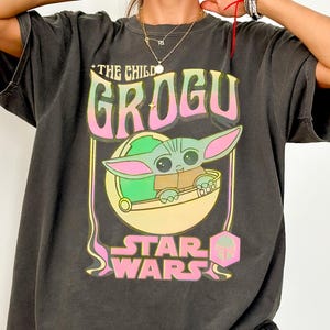 May include: Dark grey t-shirt featuring a Grogu graphic from Star Wars. The design includes the text "THE CHILD GROGU" in pink and yellow, with an image of Grogu in a pod. The words "STAR WARS" are below the image. The shirt is a Comfort Colors Tee.