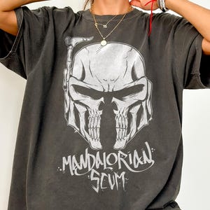 Pu&ograve; includere: T-shirt Comfort Colors grigio scuro con grafica bianca di un casco a forma di teschio e la scritta "MANDALORIAN SCUM". La maglietta oversize ha maniche corte. Il tessuto &egrave; morbido e confortevole.
