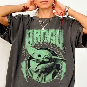 May include: Dark grey Comfort Colors tee featuring a green graphic of Grogu, with the word "GROGU" above the character. The design includes green lightning bolts and flames. The shirt is a relaxed fit.