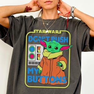 May include: Dark grey t-shirt featuring a graphic of Grogu from Star Wars with the text "DON'T PUSH MY BUTTONS". The graphic includes a green Grogu wearing an orange coat. The shirt is a Comfort Colors Tee in Pepper.