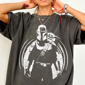 May include: Dark grey Comfort Colors tee featuring a white graphic of a Mandalorian character and Grogu. The design is set within a circular frame. The shirt has short sleeves and a relaxed fit. The text "Comfort Colors Tee (PEPPER)" is printed at the bottom.
