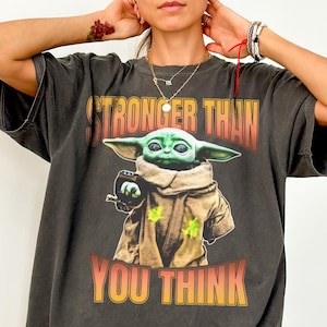 Pode incluir: T-shirt cinza escuro Comfort Colors com um gr&aacute;fico de Grogu de The Mandalorian. A imagem mostra Grogu com o texto "STRONGER THAN YOU THINK" em laranja e amarelo. A camisa tem um ajuste folgado.