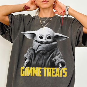 Op de afbeelding: Donkergrijs Comfort Colors T-shirt met een afbeelding van een personage met grote oren en de tekst "GIMME TREATS" in het geel. Het T-shirt heeft een relaxte pasvorm.