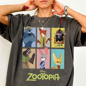 May include: Dark gray Comfort Colors tee featuring a colorful graphic of characters from the Disney movie Zootopia. The shirt has a crew neck and short sleeves. The word "Zootopia" is printed in green and white below the graphic.
