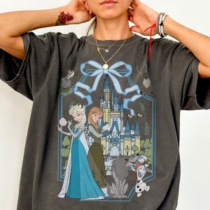 May include: Dark grey Comfort Colors tee featuring a graphic of Elsa and Anna from Frozen, with a castle backdrop and characters like Olaf and Sven. The design is framed by a blue ribbon and floral elements.