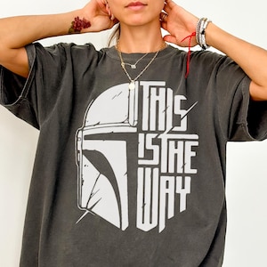 May include: Dark grey Comfort Colors tee with a white graphic of a helmet and the text "THIS IS THE WAY." The shirt has short sleeves and a relaxed fit. The text is in a bold, stylized font.