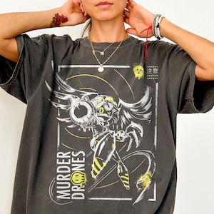 Peut inclure: T-shirt gris foncé avec un motif de personnage robotique et le texte "MURDER DRONES" en blanc et jaune. Le design comprend un symbole de risque biologique et du texte japonais. Le t-shirt est un Comfort Colors Pepper.